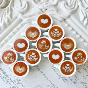 May include: A white triangular display stand with 12 small white cups filled with brown coffee. Each cup has a different design in the coffee foam, including hearts, swirls, and leaves.