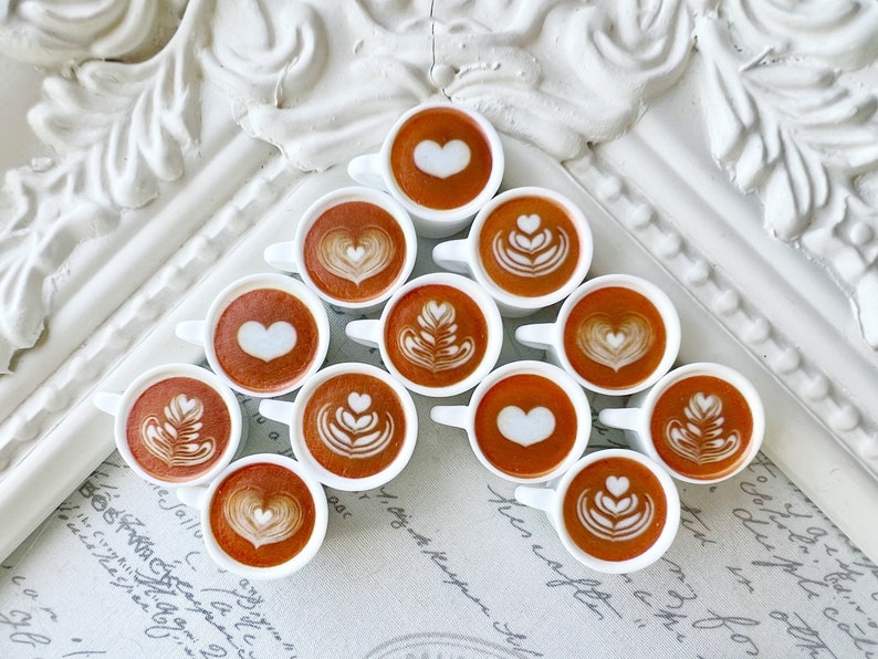 May include: A white triangular display stand with 12 small white cups filled with brown coffee. Each cup has a different design in the coffee foam, including hearts, swirls, and leaves.