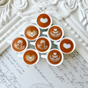 May include: Nine miniature white ceramic coffee cups with latte art designs. The cups are arranged in a triangle shape on a white and grey surface with handwritten text.