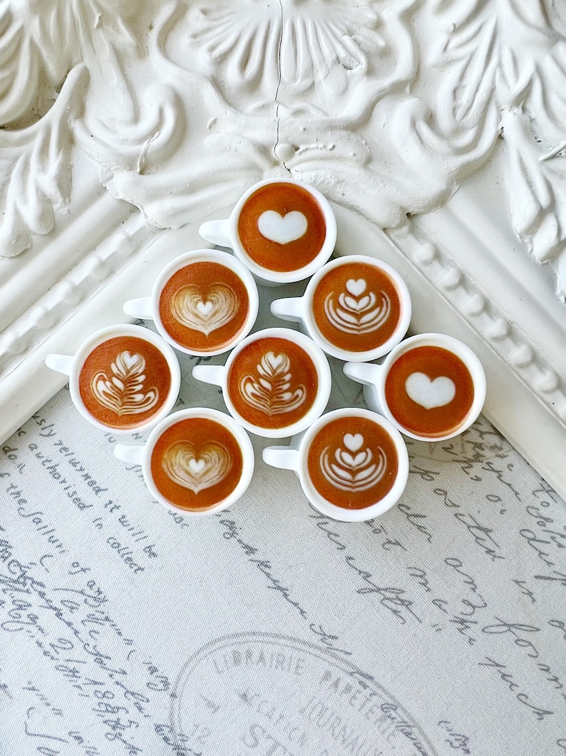 May include: Nine miniature white ceramic coffee cups with latte art designs. The cups are arranged in a triangle shape on a white and grey surface with handwritten text.