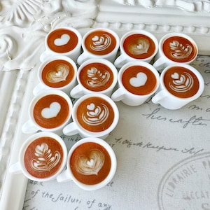 May include: A collection of 10 small white ceramic mugs filled with brown coffee with various latte art designs, including hearts, leaves, and swirls. The mugs are arranged in a staggered pattern on a white and grey surface with handwritten text.