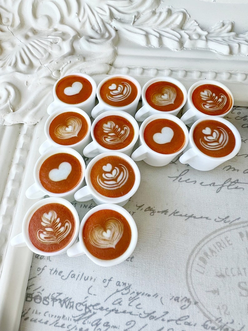 May include: A collection of 10 small white ceramic mugs filled with brown coffee with various latte art designs, including hearts, leaves, and swirls. The mugs are arranged in a staggered pattern on a white and grey surface with handwritten text.
