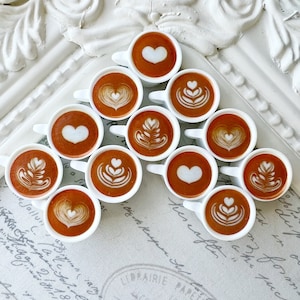 May include: A white surface with a decorative pattern is covered with a triangular arrangement of small white cups filled with coffee. Each cup has a different latte art design, including hearts, leaves, and swirls.