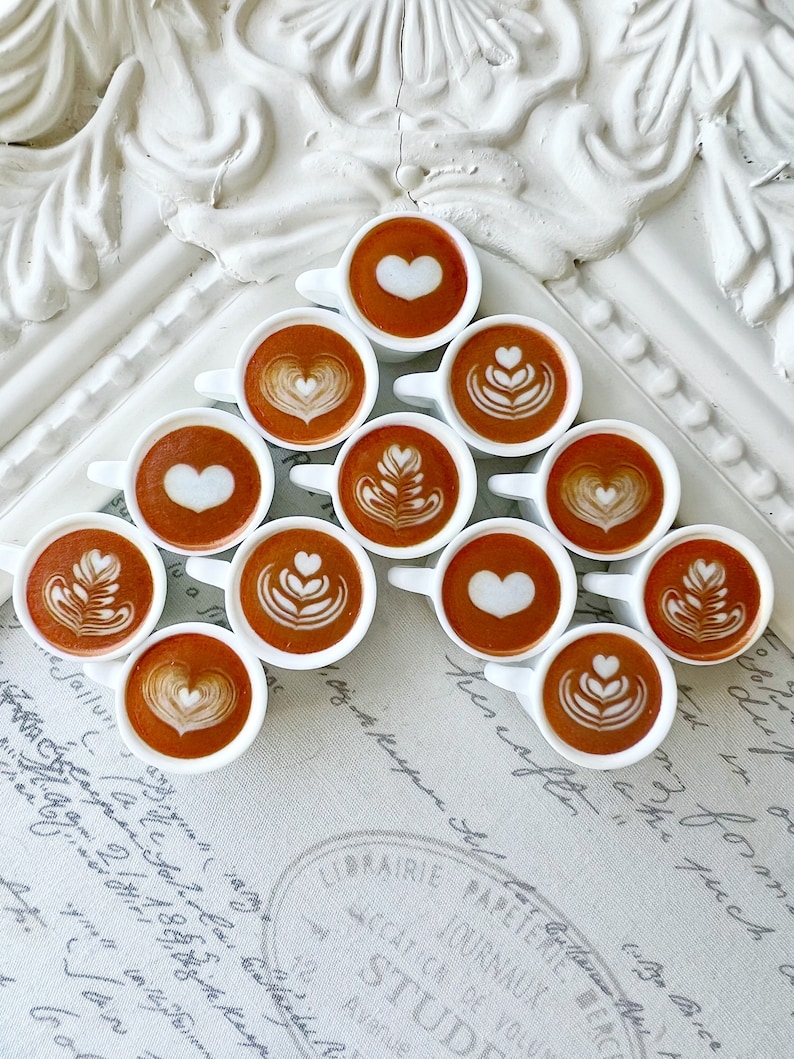 May include: A white surface with a decorative pattern is covered with a triangular arrangement of small white cups filled with coffee. Each cup has a different latte art design, including hearts, leaves, and swirls.