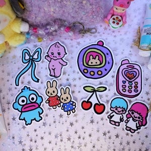 May include: A collection of colorful stickers featuring cartoon characters, a bow, and cherries. Also visible are tubes of paint, a plush toy, and a small figurine. The stickers have a kawaii aesthetic and are arranged on a star-patterned surface.
