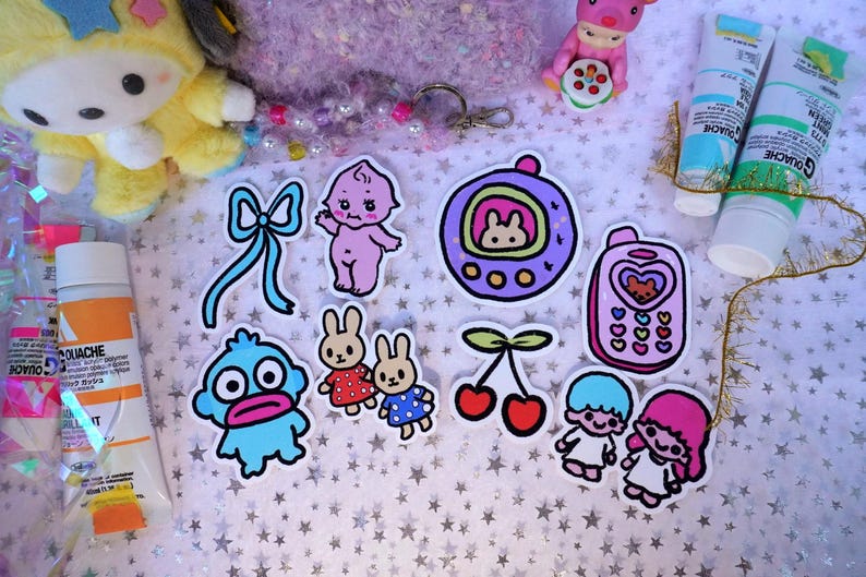 May include: A collection of colorful stickers featuring cartoon characters, a bow, and cherries. Also visible are tubes of paint, a plush toy, and a small figurine. The stickers have a kawaii aesthetic and are arranged on a star-patterned surface.