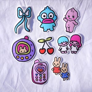 May include: A collection of colorful stickers featuring various cartoon characters and designs. Includes a blue bow, a blue creature with a pink mouth, a pink baby, a purple handheld game, red cherries, two cartoon twins, a pink phone, and two bunnies.