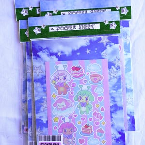 May include: Several sticker sheets in clear plastic packaging. The sticker sheets feature a variety of cute cartoon characters, cakes, and hearts. The packaging has a blue sky and star design with the text "STICKER SHEET" and "STICKER HAUL".