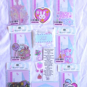 May include: A collection of stickers in various designs, including a sword, a bunny in a heart, and cartoon characters. The stickers are packaged in clear plastic with pink and blue backing cards. Text on the packaging reads "STICKER HAUL" and "I LOVE (MYSELF)".