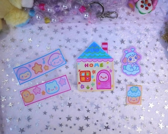 May include: A collection of colorful stickers and charms on a star-patterned surface. The stickers feature cute cartoon characters, stars, and a house with the word "HOME". Other items include a keychain and a plush toy.