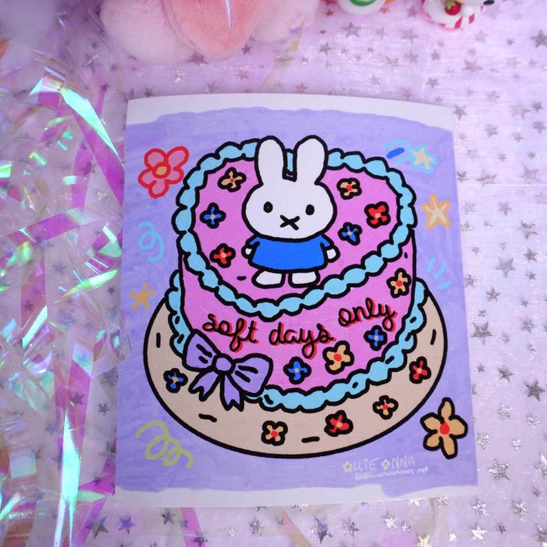May include: A sticker featuring a cartoon cake with a bunny character on top. The cake is pink with blue frosting and decorated with flowers. The text "soft days only" is written on the cake. The background is a light purple color.