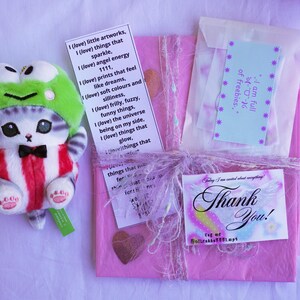May include: A plush cat toy wearing a green frog hat and a red and white striped outfit. A pink package tied with ribbon and a "Thank You" card are also visible. The image includes text on several small cards.