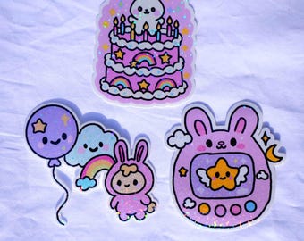 May include: Three colorful stickers with a cute theme. One sticker is a pink cake with a bunny on top. Another sticker features a bunny in a pink outfit with a rainbow and cloud. The third sticker is a pink handheld game with a star.