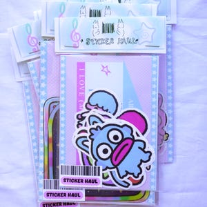 May include: A collection of sticker packs in clear plastic sleeves. The stickers feature cartoon characters, stars, and musical notes. The text "STICKER HAUL" is visible on the packaging. The color palette includes pink, blue, and purple.