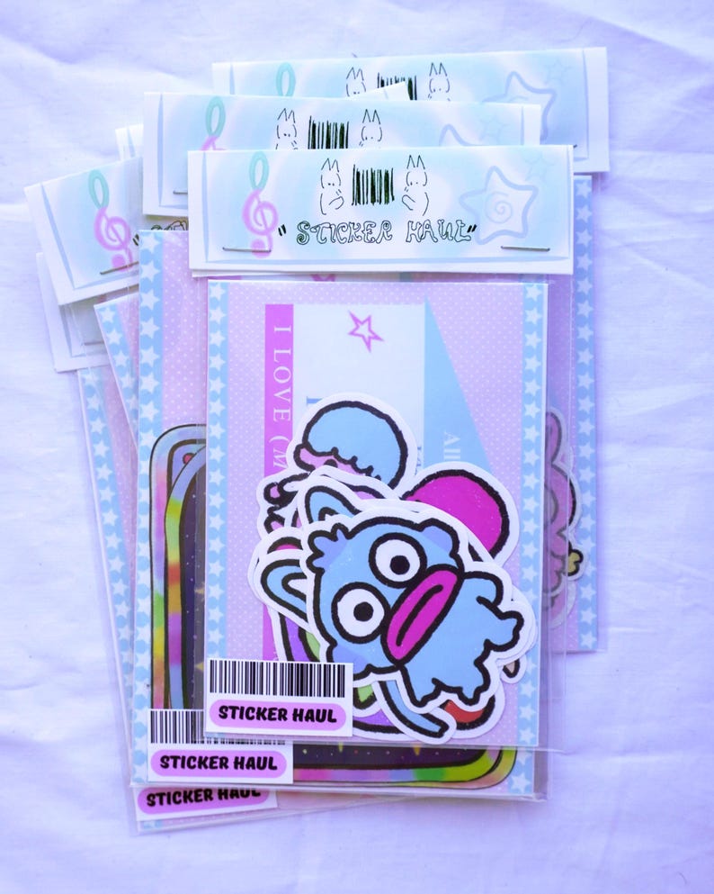 May include: A collection of sticker packs in clear plastic sleeves. The stickers feature cartoon characters, stars, and musical notes. The text "STICKER HAUL" is visible on the packaging. The color palette includes pink, blue, and purple.