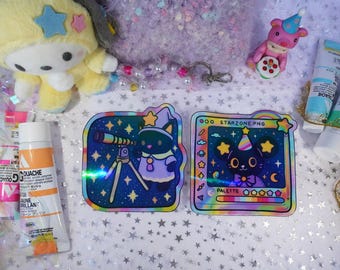May include: Two holographic stickers with cat illustrations, one with a telescope and the other with a party hat. A yellow plush toy, pink bear figurine, and tubes of paint are also visible.