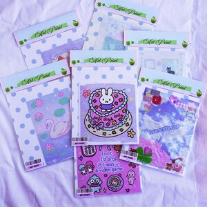 May include: A collection of art prints in clear plastic sleeves, each with a unique design. The prints feature illustrations of a swan, a cake with a bunny, and other cute graphics. The packaging includes the text "Art Print".