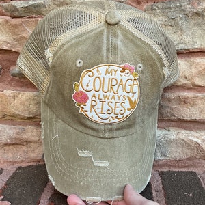 May include: A distressed olive green trucker hat with a brown mesh back. The hat has a round embroidered patch with the text "& My Courage Always Rises" in gold and white lettering with pink and yellow floral accents.