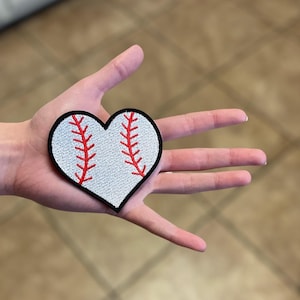 May include: A white heart-shaped patch with red stitching, resembling a baseball. The patch has a black outline.