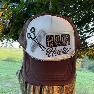May include: A brown and white trucker hat with a mesh back. The front of the hat has a white patch with a pair of scissors and a comb, and the text "HAIR Hustler" in a leopard print font.