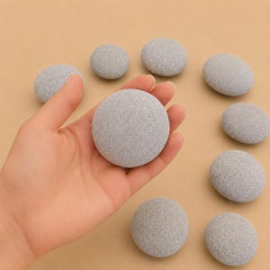 May include: A collection of smooth, round, light gray stones with a speckled texture. One stone is held in the palm of a hand, while eight other identical stones are arranged in a circular pattern around it on a solid tan surface.