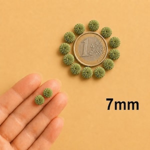 Peut inclure: Une collection de petites perles rondes en forme d'épines d'oursin vert olive, disposées en cercle autour d'une pièce d'un euro pour l'échelle. Deux perles supplémentaires sont tenues au premier plan. Le texte 7mm est imprimé en noir en bas à droite.