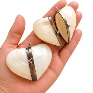 Puede incluir: Dos recipientes en forma de corazón hechos de mitades de conchas marinas de color blanco hueso e iridiscente. Cada concha presenta un marco metálico oscuro con bisagras y un pequeño cierre de bola, lo que permite abrir y cerrar los recipientes.