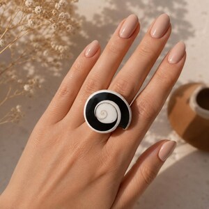 Puede incluir: Un anillo con un diseño circular de cara plana y un patrón de espiral en blanco y negro de alto contraste. El centro de la espiral es blanco, rodeado por una banda negra gruesa que se curva hacia afuera formando una figura de concha.