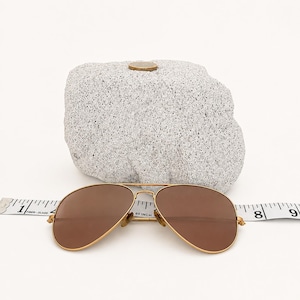 Peut inclure: Lunettes de soleil aviateur dorées avec des verres marron placées devant une pierre grise tachetée. Un ruban à mesurer blanc est placé derrière les lunettes, indiquant une largeur d'environ 14 cm d'une charnière à l'autre.
