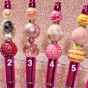 May include: Six decorative pens with pink metallic barrels and various beaded tops. The tops feature a variety of colors and designs, including rhinestones, pearls, and patterned beads. Each pen is numbered from 1 to 6.