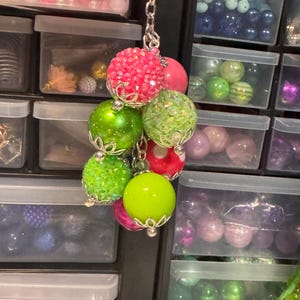 May include: A keychain featuring a cluster of colorful beads in shades of pink, green, and red. The beads have a variety of textures, including smooth, glittery, and textured surfaces. The keychain is attached to a silver-colored chain.