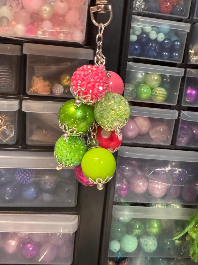 May include: A keychain featuring a cluster of colorful beads in shades of pink, green, and red. The beads have a variety of textures, including smooth, glittery, and textured surfaces. The keychain is attached to a silver-colored chain.