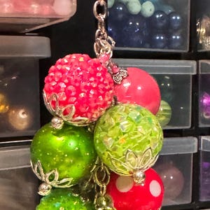 May include: A beaded keychain featuring a cluster of colorful, decorative beads. The beads are various shades of red, green, and pink, with silver-colored metal accents. Some beads have a textured, sparkly surface.