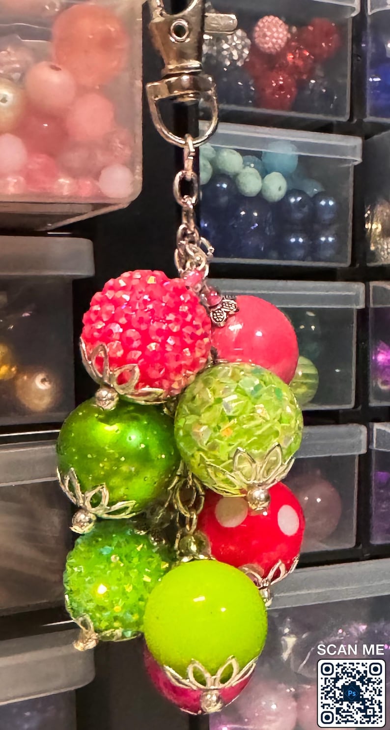 May include: A beaded keychain featuring a cluster of colorful, decorative beads. The beads are various shades of red, green, and pink, with silver-colored metal accents. Some beads have a textured, sparkly surface.