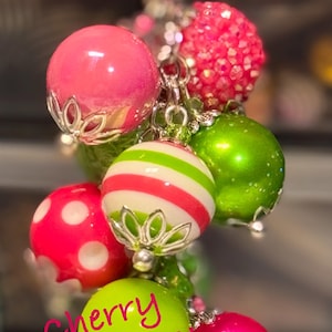 May include: A cluster of colorful, decorative baubles in shades of pink, green, and white, suspended from a silver chain. Some baubles have polka dots or stripes. The text "Cherry Limeade!" is written in pink cursive.