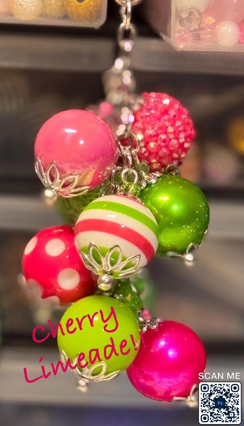 May include: A cluster of colorful, decorative baubles in shades of pink, green, and white, suspended from a silver chain. Some baubles have polka dots or stripes. The text "Cherry Limeade!" is written in pink cursive.
