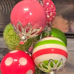 May include: A cluster of colorful, decorative ornaments. The ornaments are various colors, including pink, green, and red, with silver-colored metal accents. Some have stripes or polka dots.