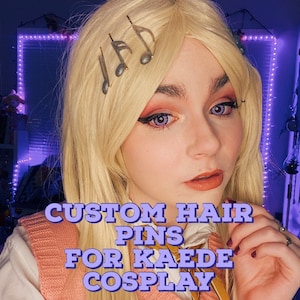 Puede incluir: Pasadores de pelo personalizados con forma de notas musicales, diseñados para un cosplay de Kaede. Los pasadores son plateados y están en el pelo rubio de una persona. La persona lleva un chaleco de punto rosa sobre una camisa blanca con una bufanda de estampado amarillo.