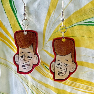 May include: A pair of earrings featuring cartoon character faces painted on wood. The characters have brown hair, brown eyes, and a light brown skin tone. The earrings are attached to silver hooks.