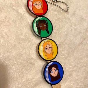 May include: A keychain featuring four circular portraits of women with different hair colors and styles. Each portrait is set against a colored background: red, green, yellow, and blue. The keychain has a silver chain and a wooden stick.