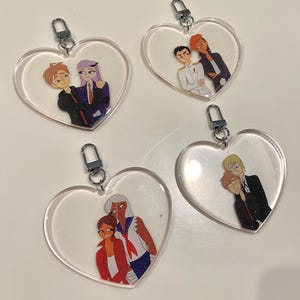 Puede incluir: Cuatro llaveros en forma de corazón con ilustraciones de parejas de dibujos animados. Cada llavero tiene un fondo transparente y brillante y un cierre plateado. Las parejas están representadas en varios atuendos y poses.