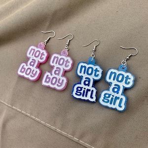 May include: Two pairs of dangle earrings with silver-toned hooks. One pair is translucent pink with white text reading not a boy. The second pair is translucent blue with white text reading not a girl. Each earring has a rounded, stacked shape.