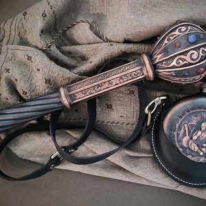 Può includere: Una fiaschetta in pelle nera con un design inciso e un cinturino in pelle marrone. La fiaschetta è attaccata a un bastone decorativo, ornato, di colore rame con una cima rotonda e decorata con gioielli.