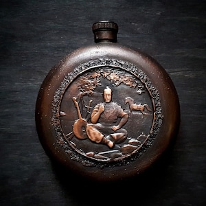 May include: A round, copper-colored flask with a detailed embossed design. The design features a man playing a musical instrument in a forest setting.