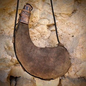 May include: A brown leather water skin with a decorative copper-coloured metal top and a black rope strap. The water skin is hanging against a textured stone wall. The leather has a worn, suede-like appearance.