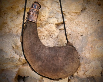 May include: A brown leather water skin with a decorative copper-colored metal top and a black rope strap. The water skin is hanging against a textured stone wall. The leather has a worn, suede-like appearance.