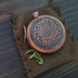 Pode incluir: Uma garrafa redonda, cor de cobre, com um desenho em relevo, pousada numa bolsa castanha. A garrafa apresenta um padrão circular detalhado. A bolsa é feita de um material castanho escuro texturizado. O fundo é uma superfície de madeira escura.