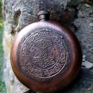 May include: A copper-toned, circular hip flask with a screw-top lid. The flask is decorated with an embossed image of Thor's hammer, framed by Celtic knot patterns. The metal flask has a vintage or antique aesthetic.