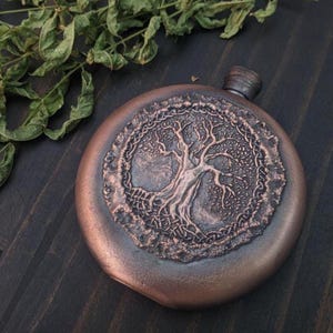 Pode incluir: Uma garrafa redonda, cor de cobre, com um design detalhado da árvore da vida no centro. A árvore é rodeada por uma borda intrincada. A garrafa tem uma pequena rolha cilíndrica e repousa sobre uma superfície de madeira escura.