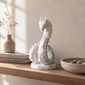 May include: A white, geometric dragon sculpture with a faceted, angular surface texture. The dragon is depicted in a coiled, upright position, featuring sharp spikes along its back and a detailed head, all mounted on a circular base.